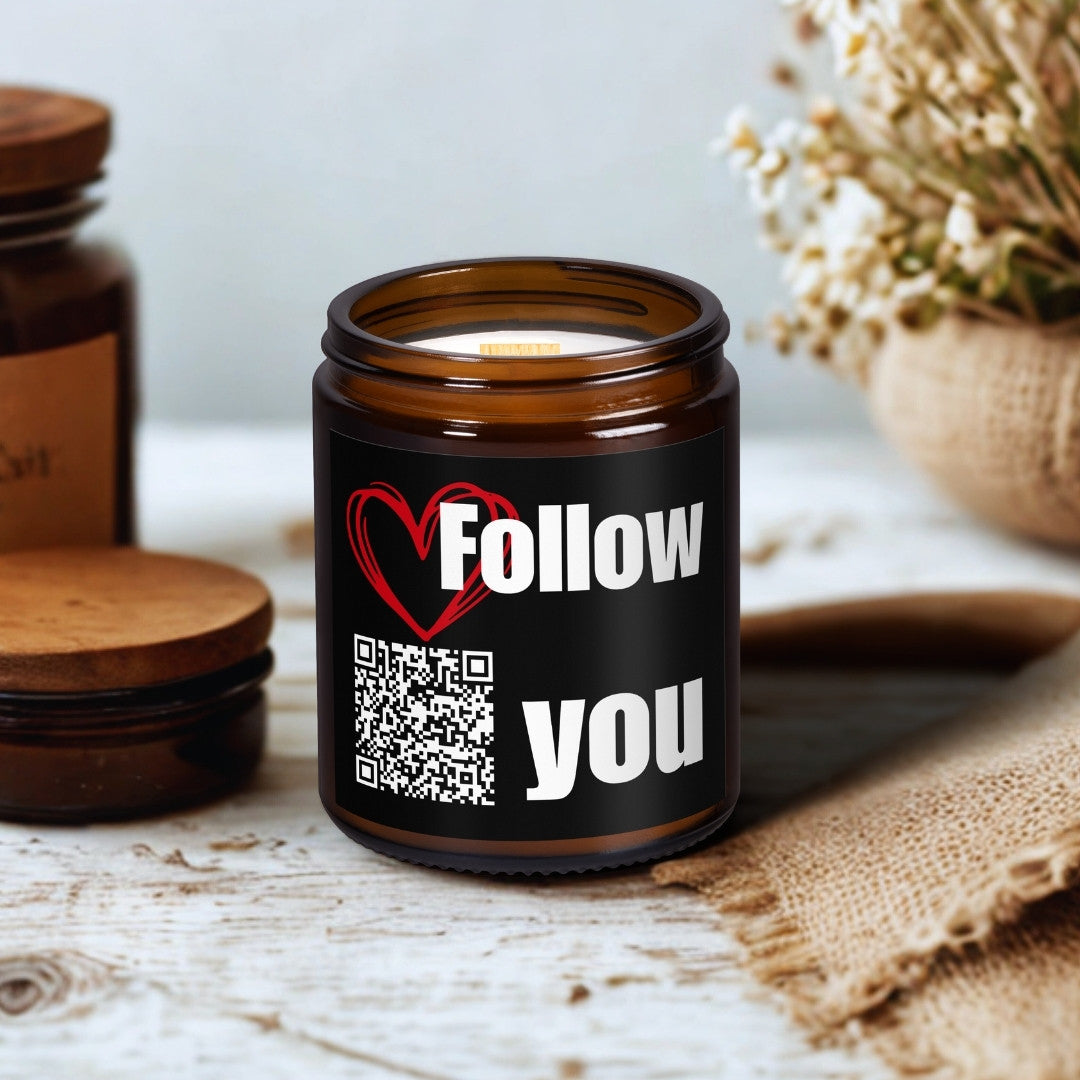 Lumânare din ceară de soia Kiskedves în borcan maro de sticlă, cu fitil de lemn; pe etichetă un cod QR și melodia Follow You, pe o masă de lemn lângă flori uscate