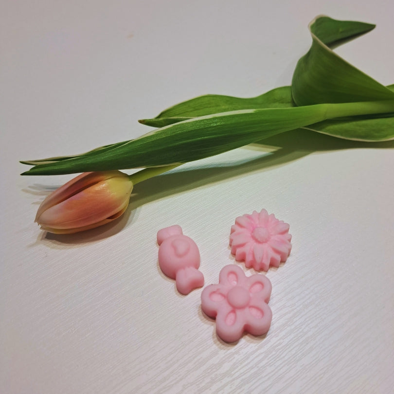 Trei bucăți de ceară parfumată Kiskedves roz – iepuraș, margaretă și floare – lângă o lalea, pe o suprafață albă de lemn.