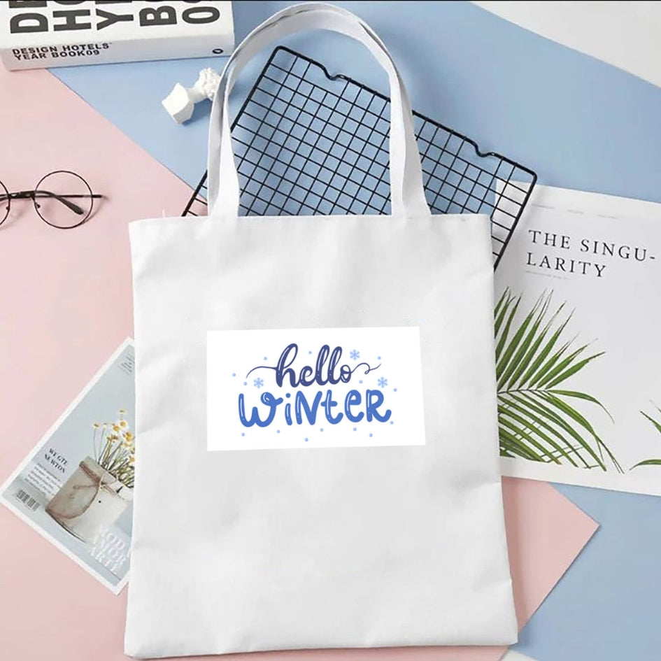Sacoșă de pânză Kiskedves Hello Winter în alb, cu text albastru hello winter, pe fundal pastel lângă reviste și ochelari.