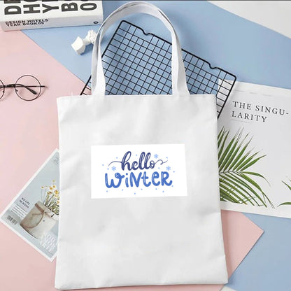 Sacoșă de pânză Kiskedves Hello Winter în alb, cu text albastru hello winter, pe fundal pastel lângă reviste și ochelari.