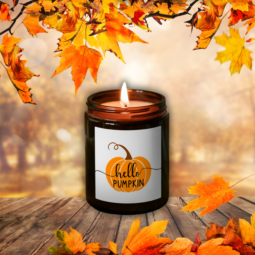 Lumânare Pumpkin Spice aprinsă, în borcan maro, cu eticheta „hello PUMPKIN”, printre frunze de toamnă pe o masă de lemn