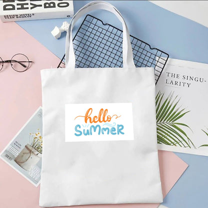 Sacoșă de pânză Kiskedves Hello Summer în alb, cu text portocaliu și albastru hello summer, pe fundal pastel lângă reviste.