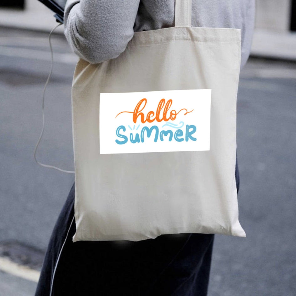 Sacoșă de pânză naturală Kiskedves Hello Summer purtată pe umăr, cu text portocaliu și albastru hello summer, pe stradă.