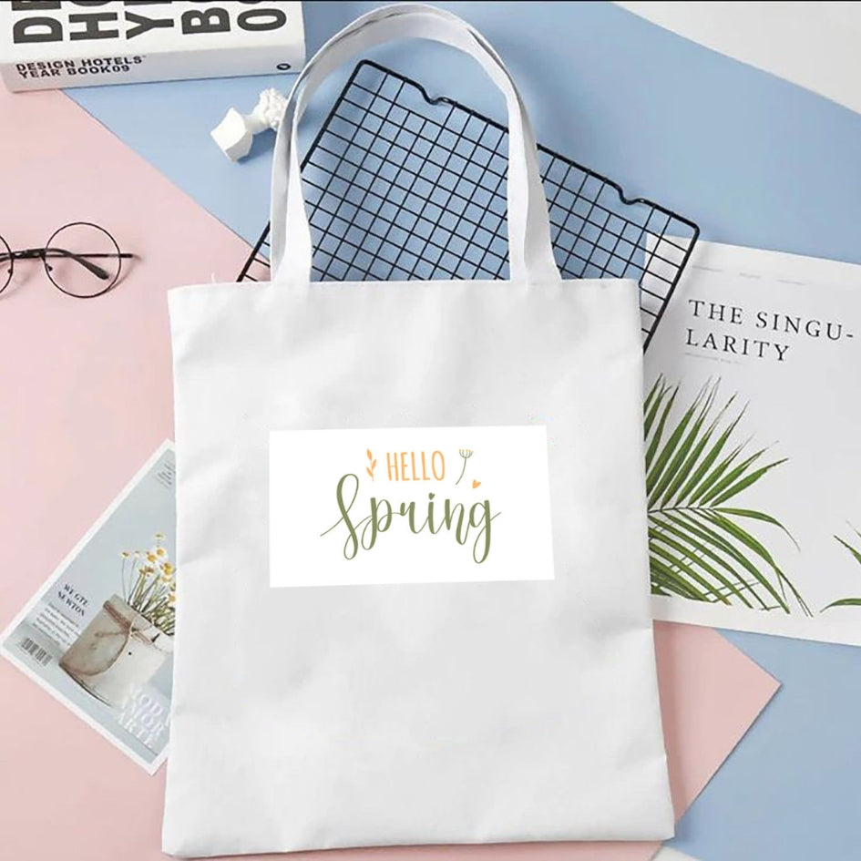 Sacoșă de pânză Kiskedves Hello Spring în alb, cu text verde și portocaliu hello spring, pe fundal pastel lângă reviste.