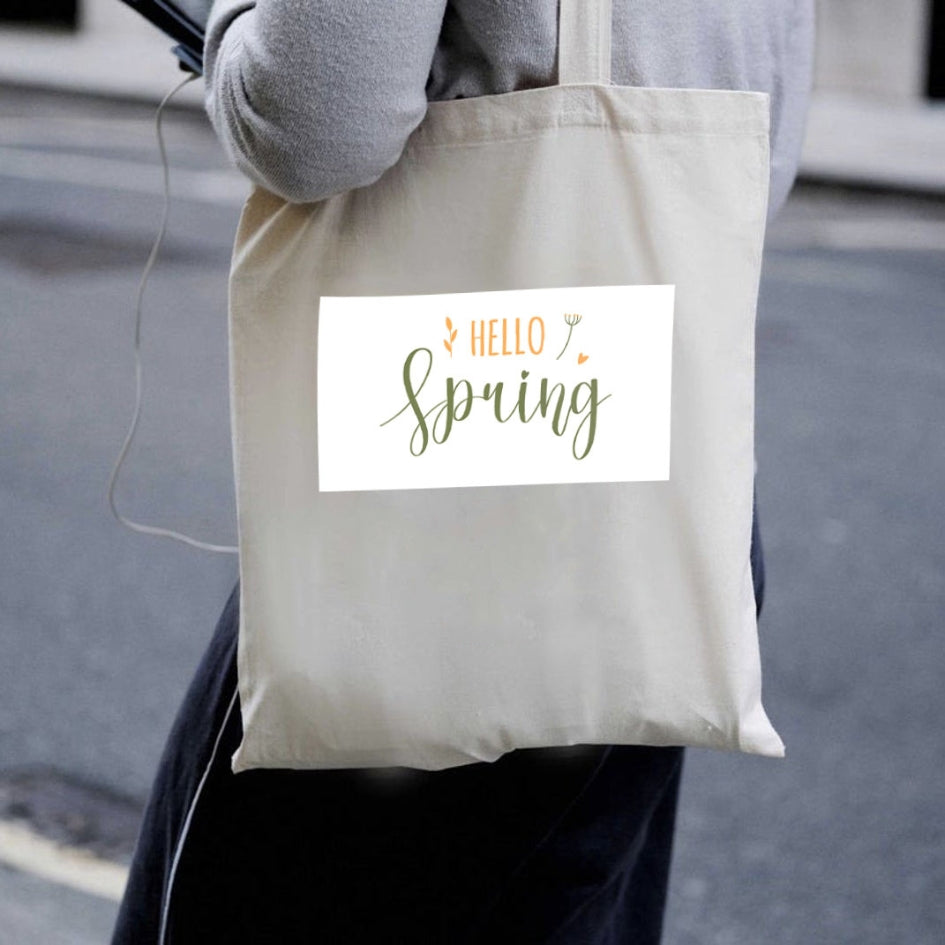 Sacoșă de pânză naturală Kiskedves Hello Spring purtată pe umăr, cu text verde și portocaliu hello spring, pe stradă.
