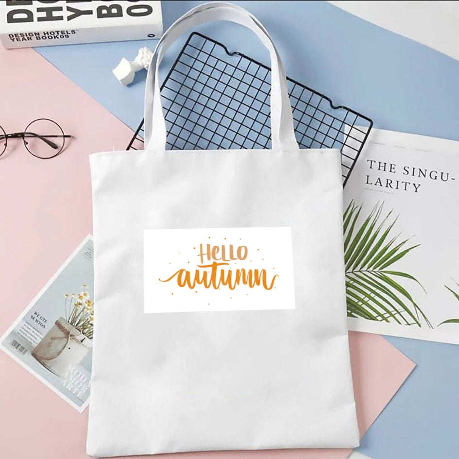 Sacoșă de pânză Kiskedves Hello Autumn în alb, cu text portocaliu hello autumn, pe fundal pastel lângă reviste.