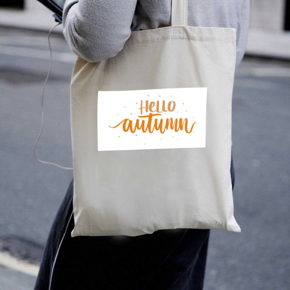Sacoșă de pânză naturală Kiskedves Hello Autumn purtată pe umăr, cu text portocaliu hello autumn, pe stradă.