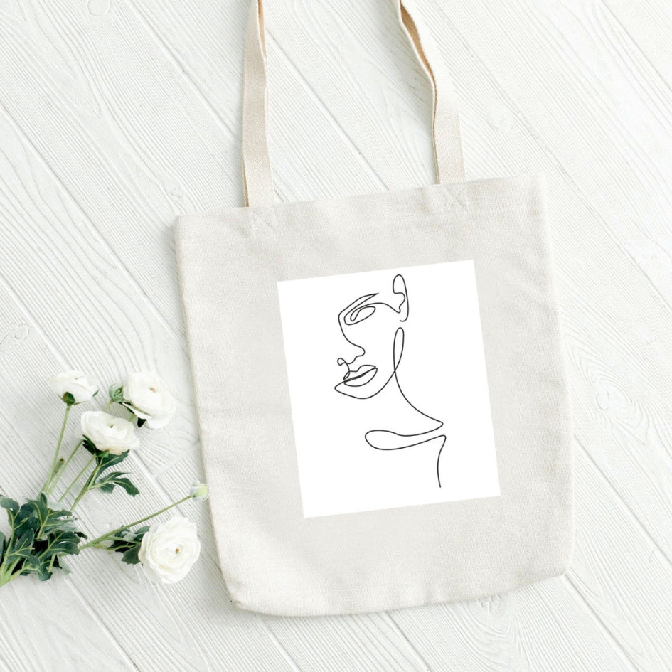 Sacoșă de pânză Kiskedves Line Art cu profil feminin, pe scânduri albe, lângă flori albe.