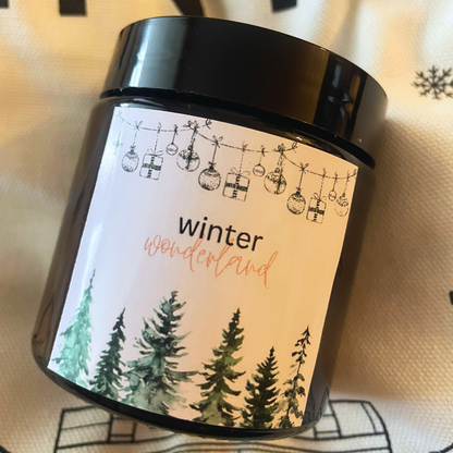Lumânare Kiskedves Winter Wonderland pe un textil cu fulgi de nea, prim-plan cu eticheta cu brazi.