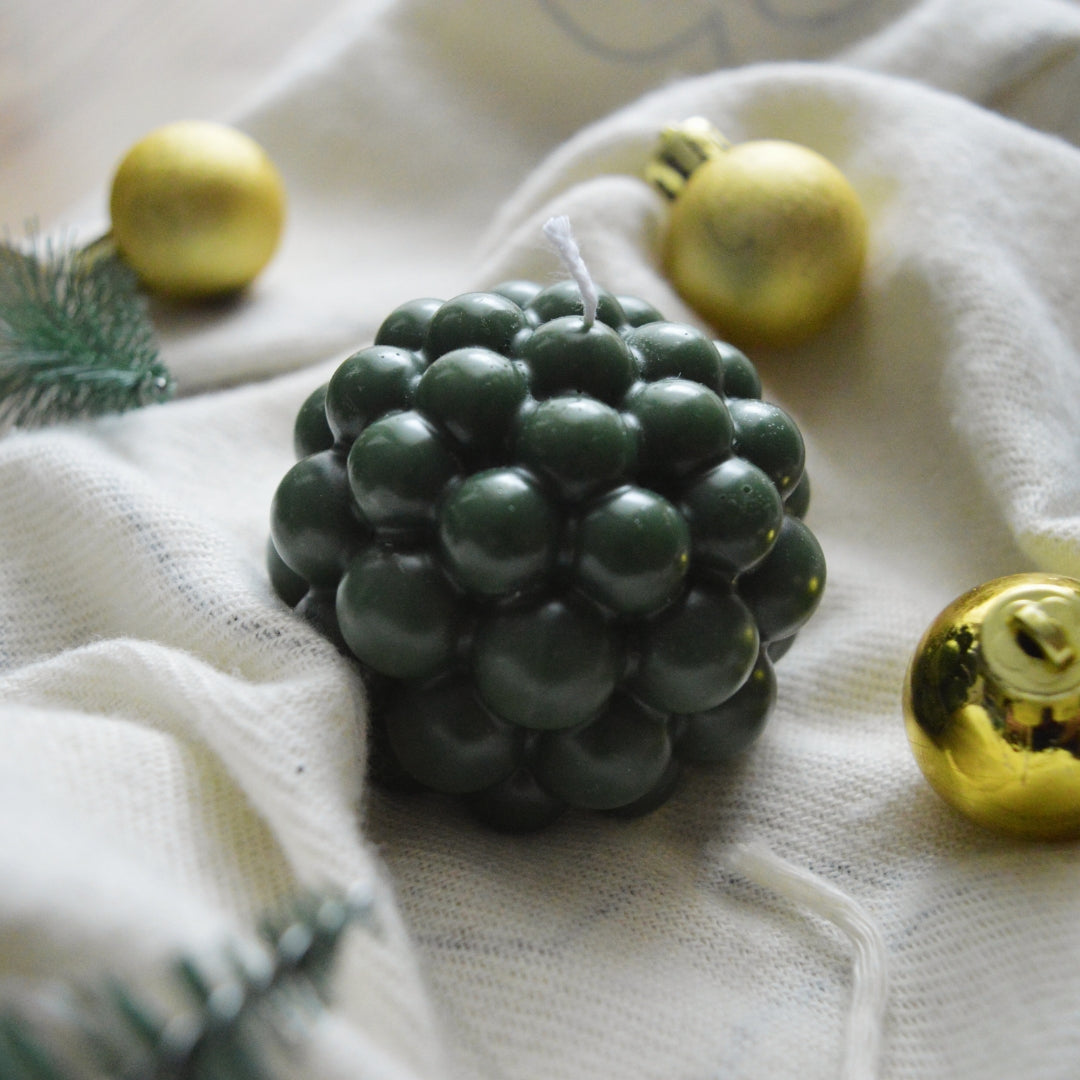 Lumânare Kiskedves Bubi verde închis pe un tricot alb, printre globuri aurii de Crăciun.