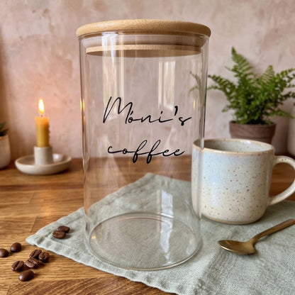 Borcan de sticlă cu text și capac de bambus, text negru, lângă o ceașcă de cafea și boabe de cafea