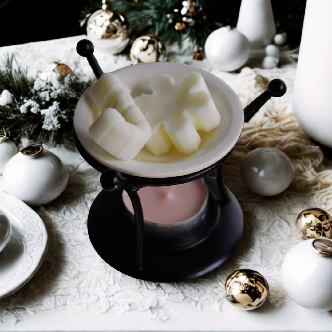 Lampă aromaterapie Kiskedves din metal negru cu bol de ceramică, în el ceruri de soia crem în forme festive, dedesubt o pastilă de ceai roz, în jur globuri albe și aurii