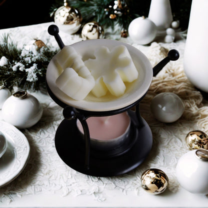 Lampă aromaterapie Kiskedves din metal negru cu bol de ceramică, în el ceruri de soia crem în forme festive, dedesubt o pastilă de ceai roz, în jur globuri albe și aurii