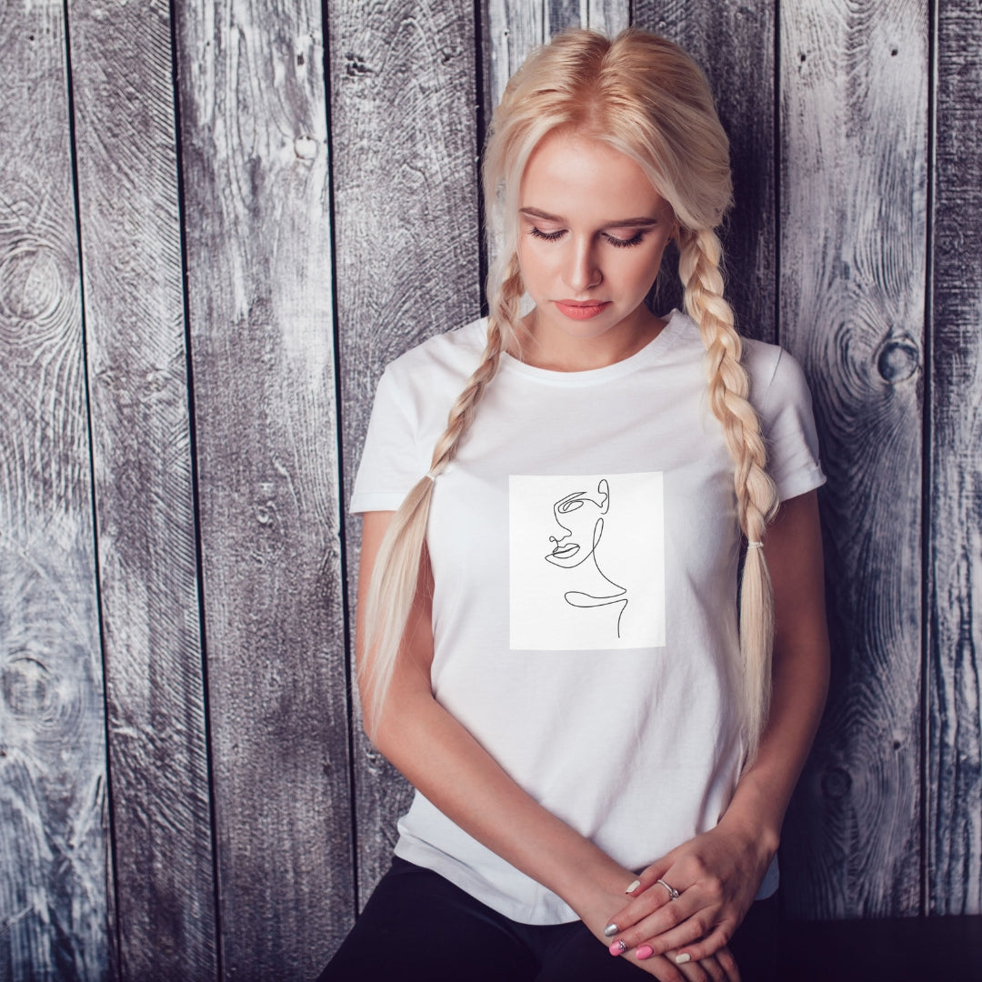 Tricou alb Kiskedves Line Art cu profil feminin, pe o modelă cu codițe, în fața unui perete de lemn.