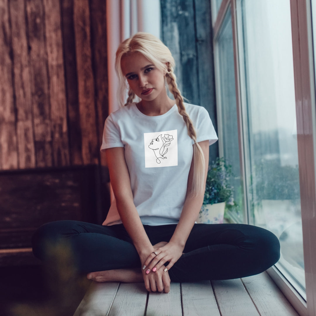 Tricou alb Kiskedves Line Art cu floare în păr, pe o modelă așezată lângă fereastră.