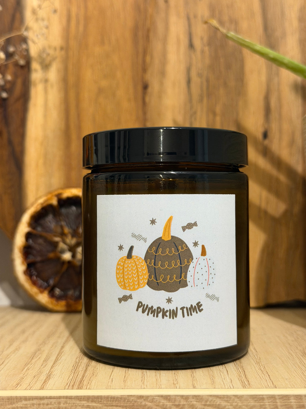 Lumânare Pumpkin Spice din ceară de soia în borcan maro, închisă cu capac negru, cu eticheta „Pumpkin time” cu dovleci desenați, pe un raft de lemn, lângă o felie de portocală uscată