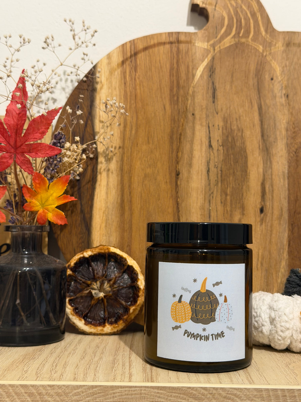 Lumânare Pumpkin Spice din ceară de soia în borcan maro, cu eticheta „Pumpkin time”, în fața unui tocător de lemn în formă de dovleac, lângă o vază cu flori uscate, frunze de toamnă și un dovleac alb croșetat