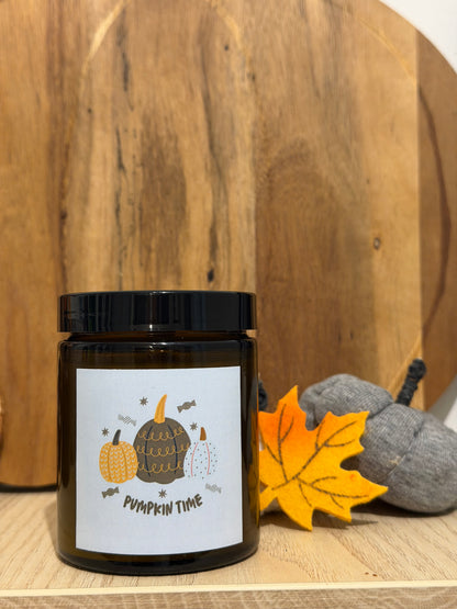 Lumânare Pumpkin Spice din ceară de soia în borcan maro, cu eticheta „Pumpkin time”, în fața unui platou rotund de lemn, lângă o frunză de arțar din fetru galben și dovleci gri din material textil
