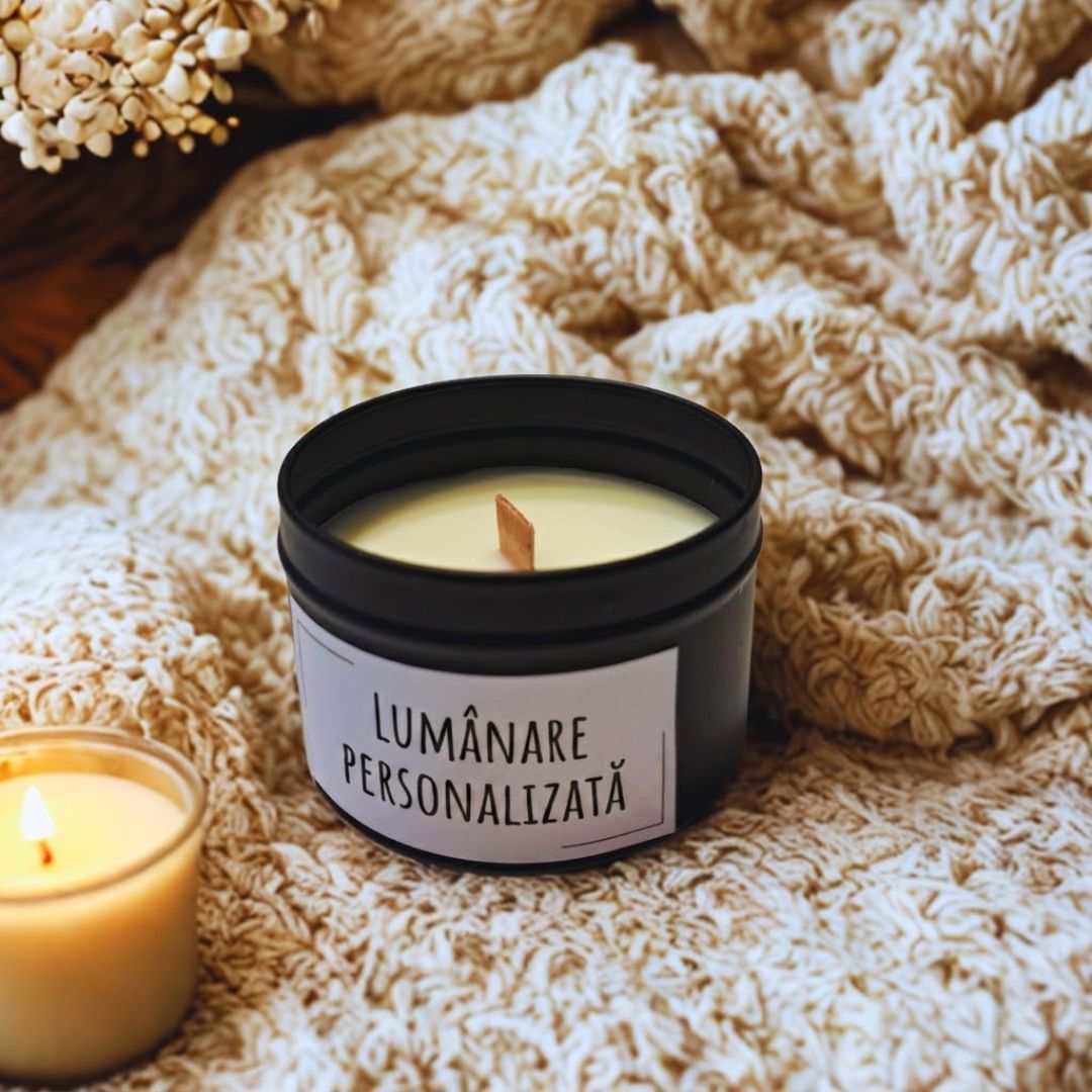 Lumânare cu fitil de lemn în cutie metalică neagră cu inscripția „Lumânare personalizată”, pe o pătură tricotată lângă o lumânare aprinsă
