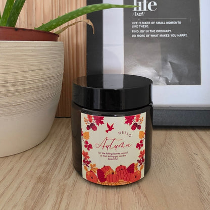 Lumânare Pumpkin Spice din ceară de soia în borcan maro, închisă cu capac negru, cu eticheta „Hello Autumn” cu o coroniță de flori de toamnă, pe un raft de lemn, lângă un aloe vera în ghiveci și un tablou înrămat