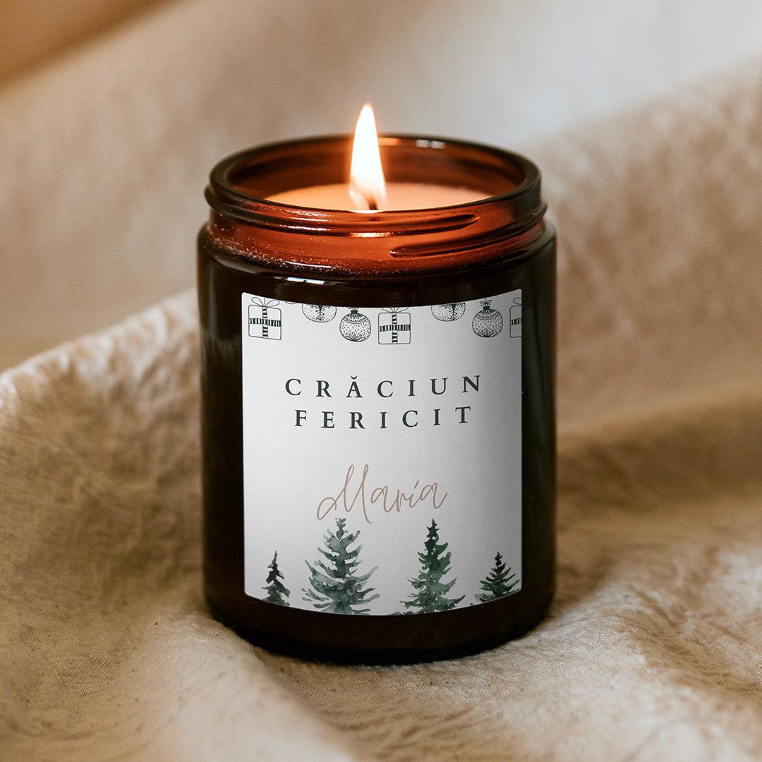 Lumânare parfumată Kiskedves aprinsă în borcan maro pe o țesătură moale, pe etichetă o ghirlandă de Crăciun, pădure în acuarelă și inscripția Crăciun fericit cu numele dorit