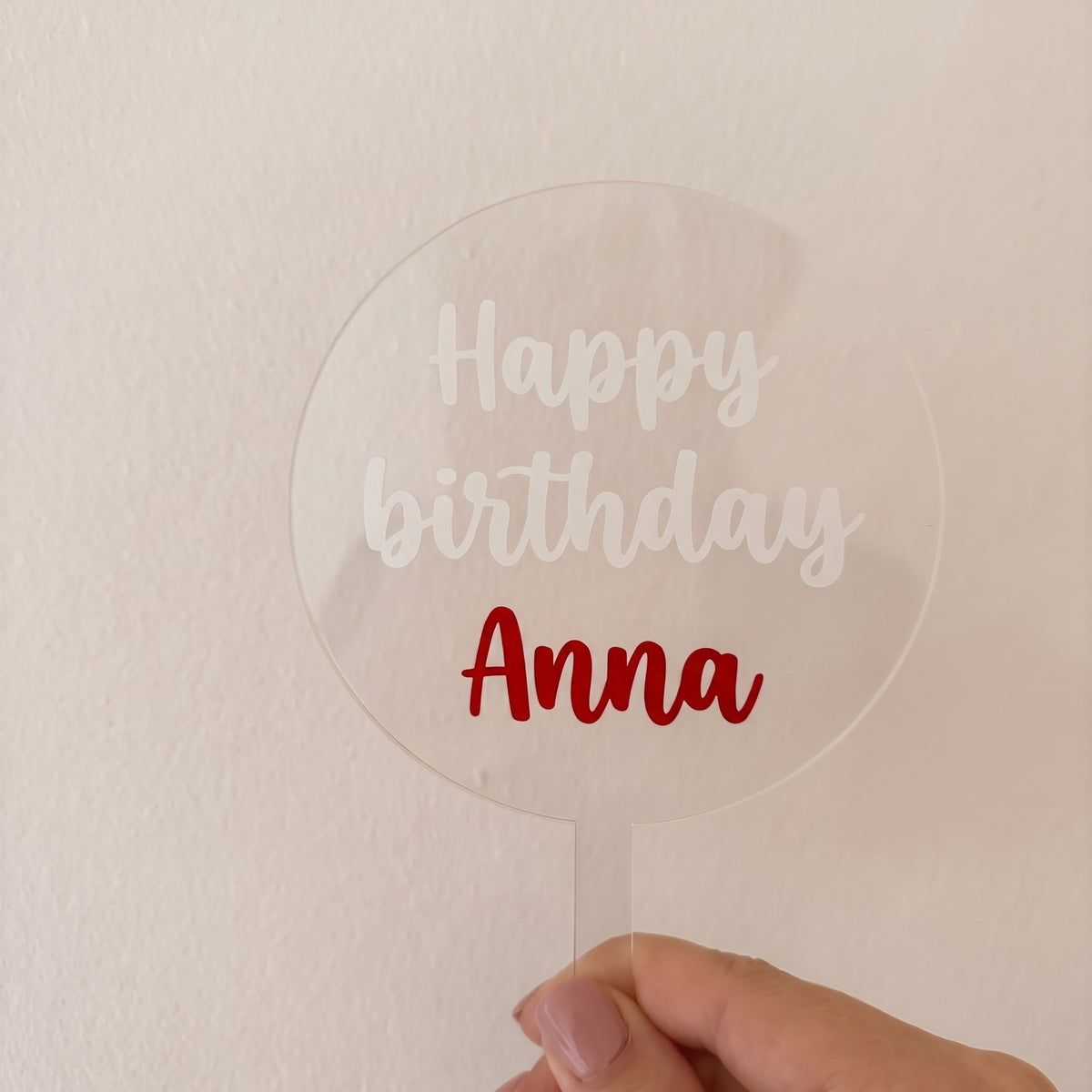 Topper transparent din acril cu textul alb „Happy birthday” și numele roșu „Anna”, ținut în mână
