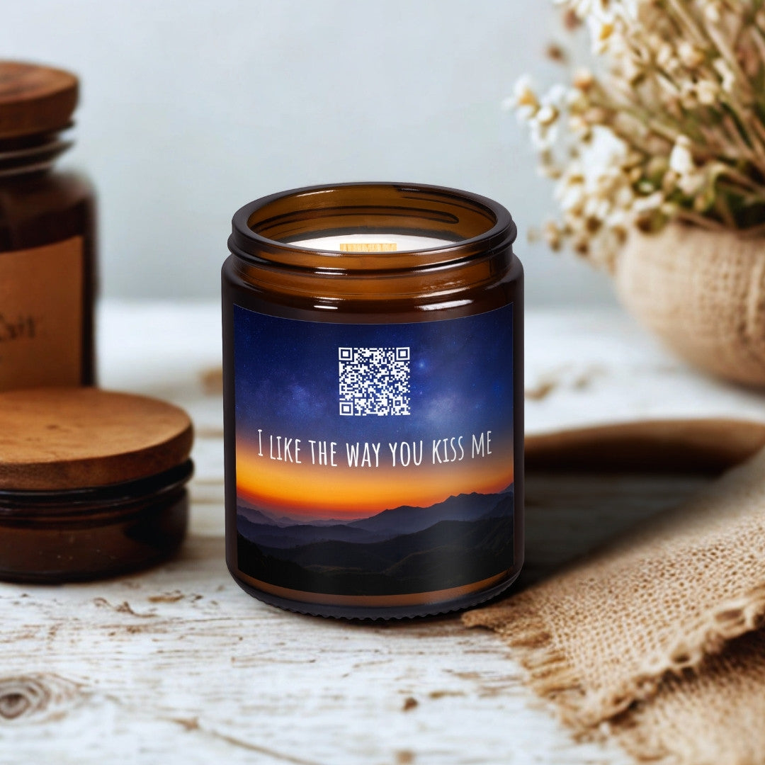 Lumânare din ceară de soia Kiskedves în borcan maro de sticlă, cu fitil de lemn; pe etichetă un cod QR și melodia I like the way you kiss me, pe o masă de lemn lângă flori uscate