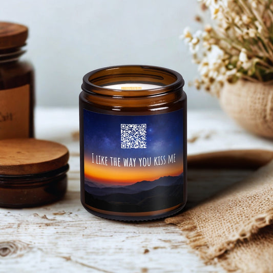 Lumânare din ceară de soia Kiskedves în borcan maro de sticlă, cu fitil de lemn; pe etichetă un cod QR și melodia I like the way you kiss me, pe o masă de lemn lângă flori uscate
