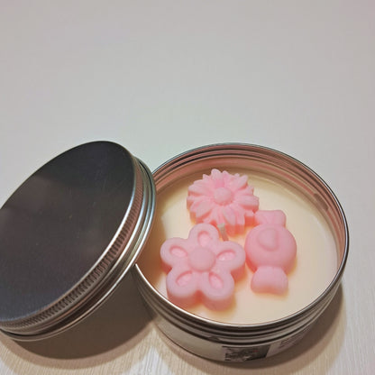 Lumânare de primăvară Kiskedves în cutie metalică argintie, cu capacul dat la o parte și floricele roz din ceară