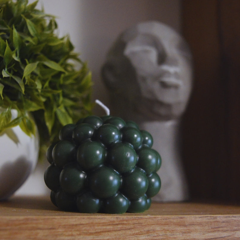 Lumânare Kiskedves Bubi verde închis cu bule, prim-plan, cu o sculptură de piatră în fundal.