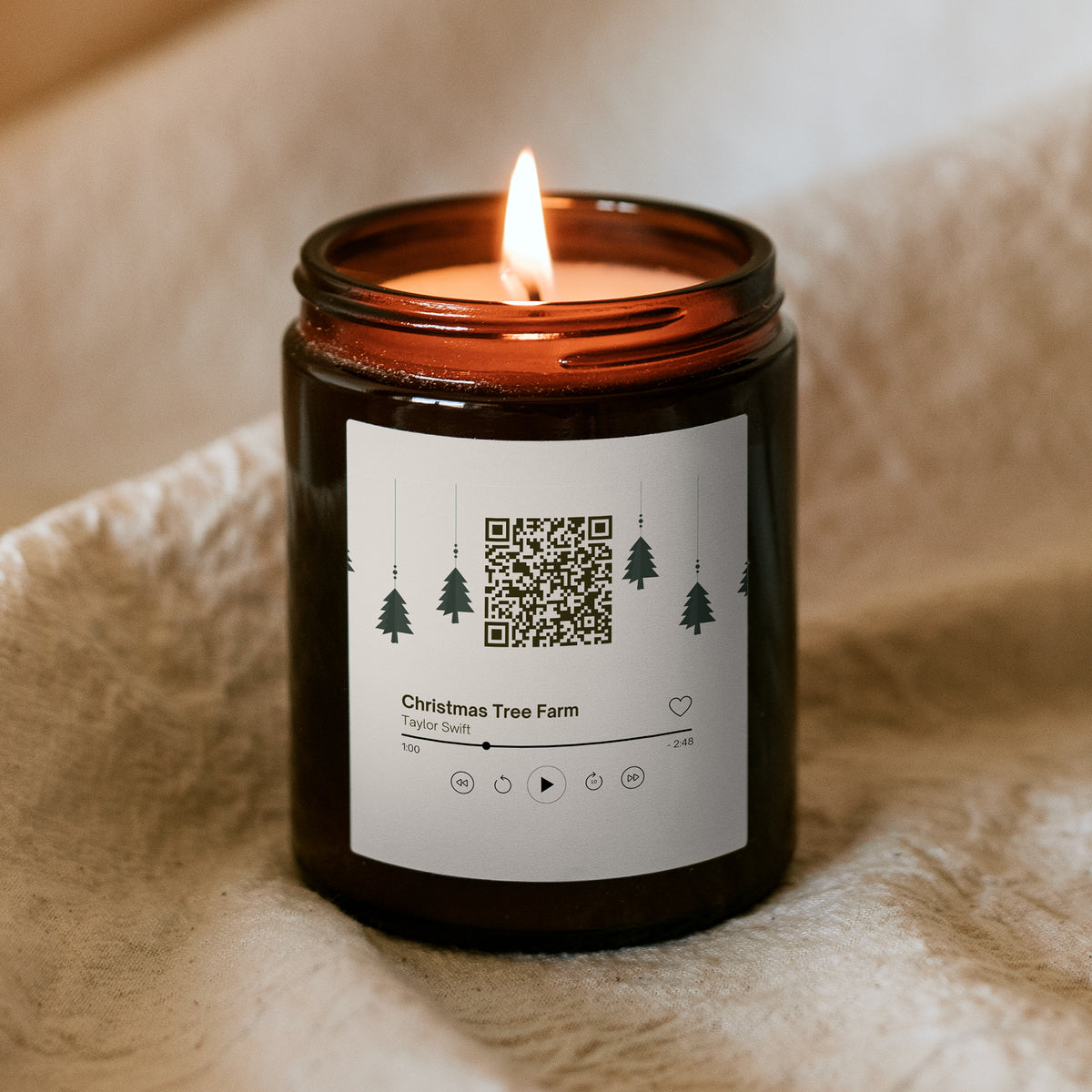 Lumânare de Crăciun „Melodică” Kiskedves aprinsă în borcan maro, pe o pătură moale, pe etichetă cod QR și un card de player cu melodia Christmas Tree Farm