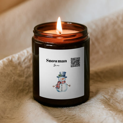 Lumânare de Crăciun „Melodică” Kiskedves aprinsă în borcan maro, pe o pătură moale, pe etichetă cod QR, inscripția Snowman și un om de zăpadă în acuarelă