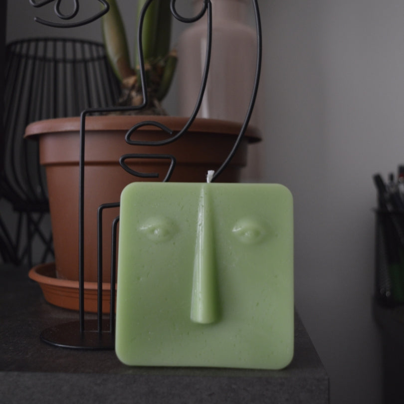 Lumânare pătrată verde cu chip Line Art pe o masă închisă la culoare, în spate o decorațiune din sârmă în formă de chip și o plantă în ghiveci