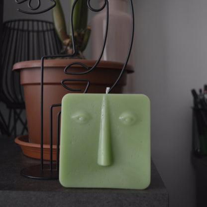 Lumânare pătrată verde cu chip Line Art pe o masă închisă la culoare, în spate o decorațiune din sârmă în formă de chip și o plantă în ghiveci