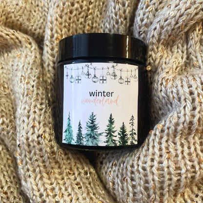 Lumânare Kiskedves Winter Wonderland pe o pătură tricotată, se vede bine eticheta acuarelă cu brazi.